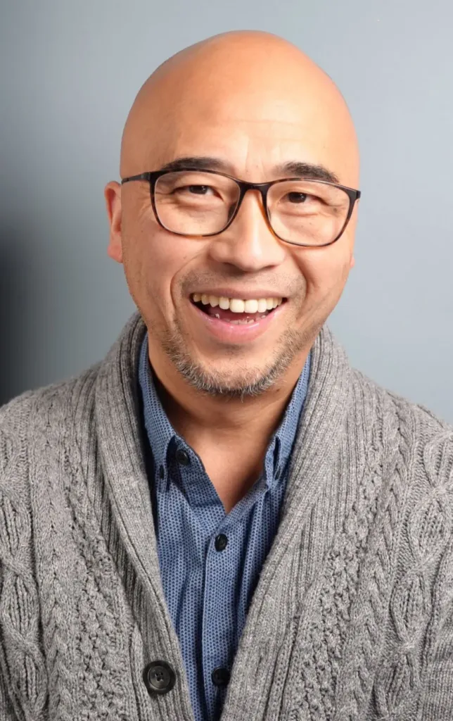 A smiling man with glasses, a shaven head, and a short beard wears a blue shirt and a grey knitted cardigan against a plain light blue background.
