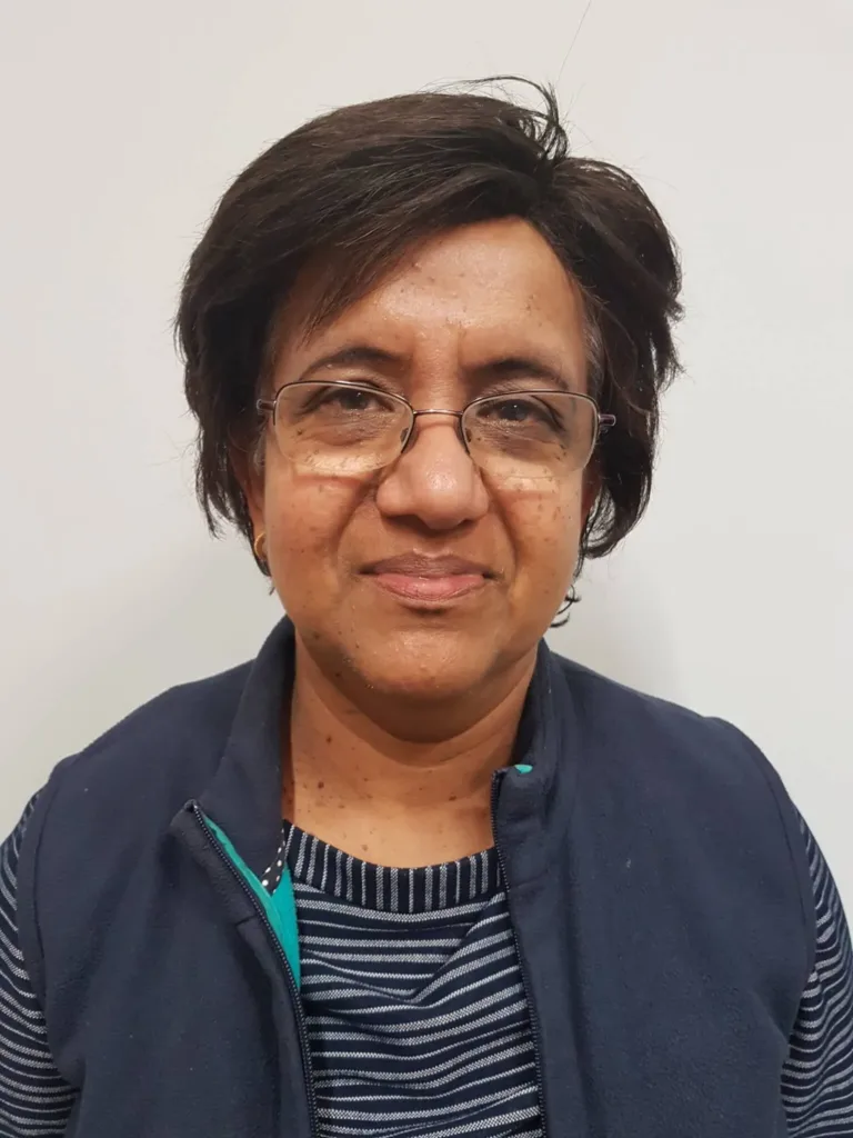 A woman with short dark hair and glasses is wearing a navy waistcoat over a striped shirt, standing against a plain white background.