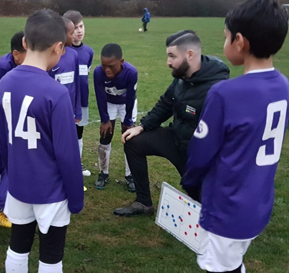 A coach kneeling with the school football team around a tactics board on the field