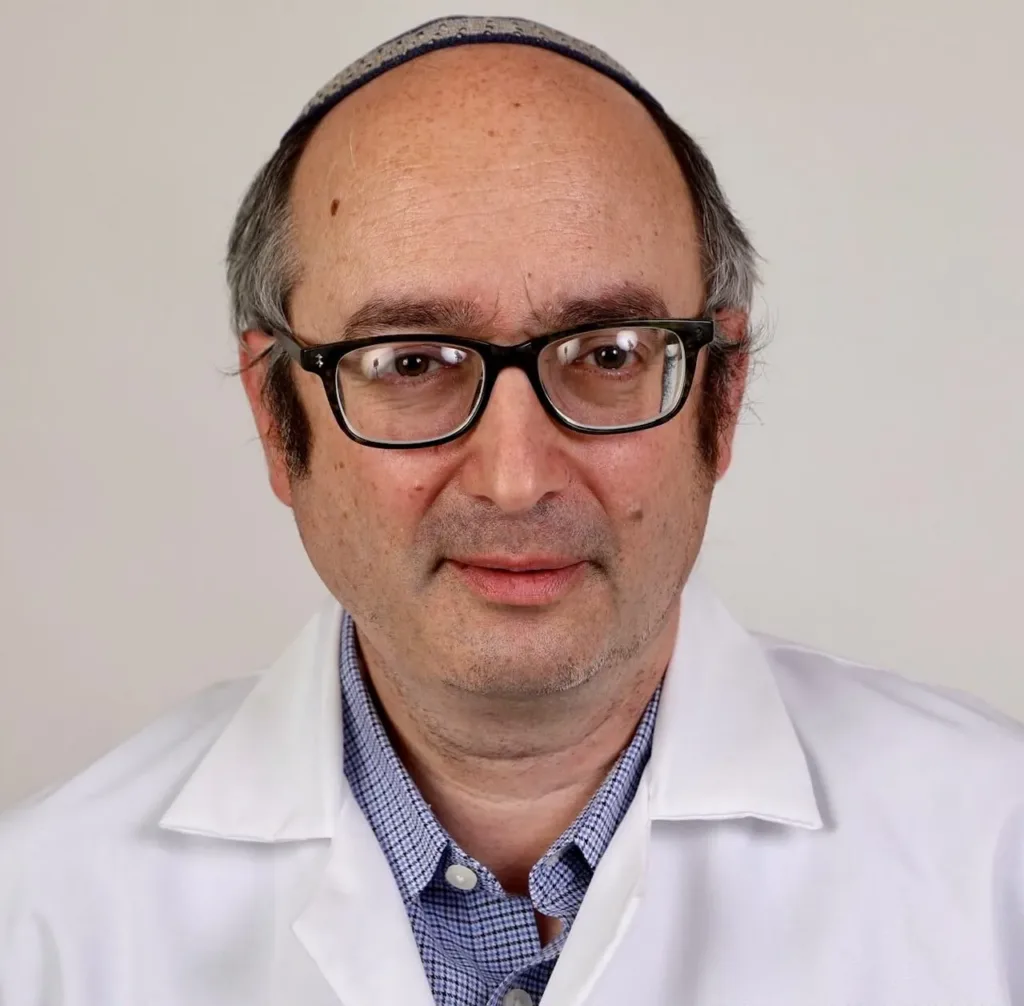 A middle-aged man wearing glasses, a blue chequered shirt, a white lab coat, and a kippah, looking at the camera with a neutral expression against a plain background.