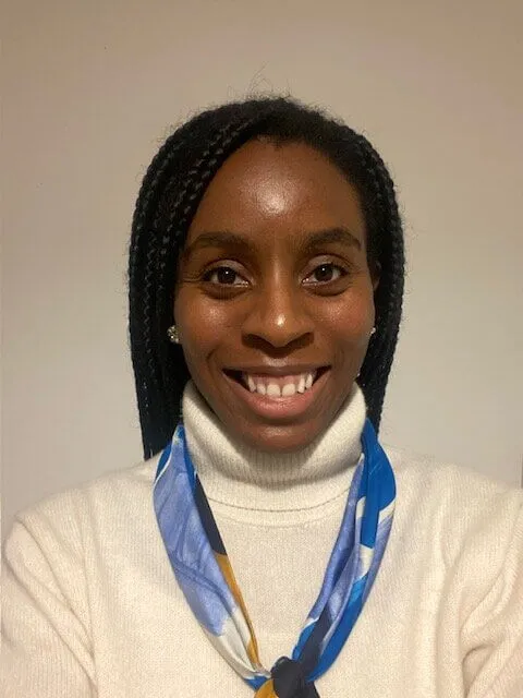 A woman with plaited hair smiles at the camera. She is wearing a white polo-neck jumper and a blue patterned scarf, standing against a plain light-coloured background.