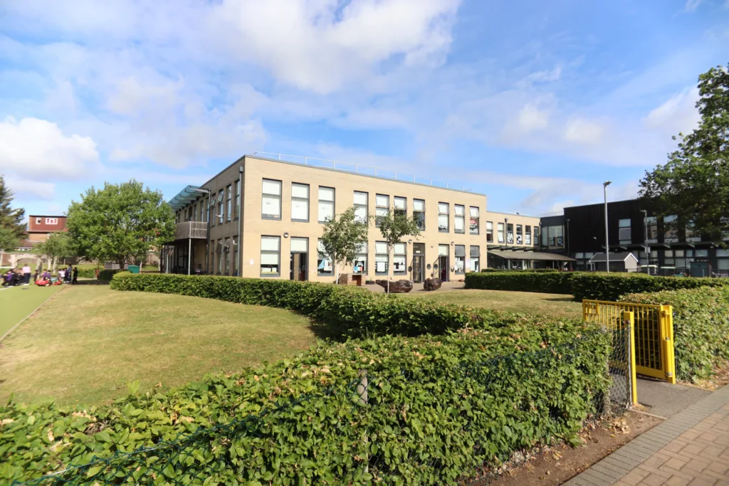 A modern, two-storey school building with large windows, surrounded by green lawns and hedges, under a partly cloudy blue sky. A yellow gate and pavement are visible in the foreground.