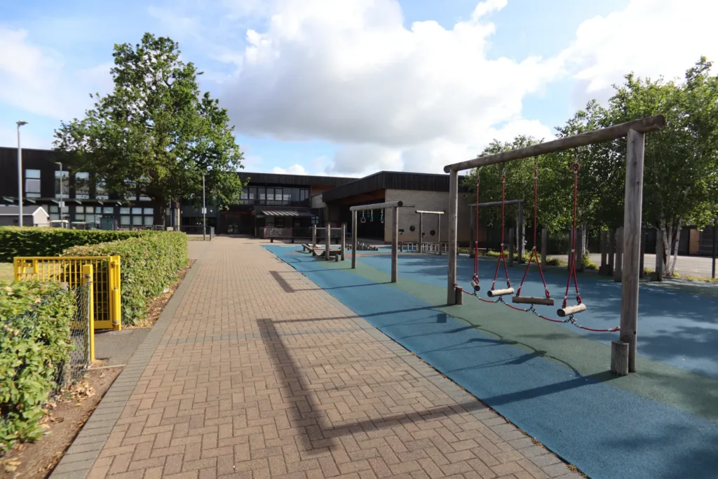 A quiet playground with swings and benches on blue flooring, bordered by hedges and trees, sits next to a modern building under a partly cloudy sky.