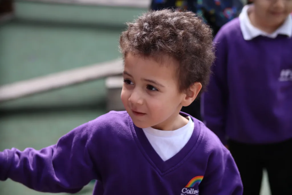 A child with curly hair wearing a purple jumper with a rainbow logo stands outdoors, looking to the side, with another child in a similar outfit partially visible in the background.