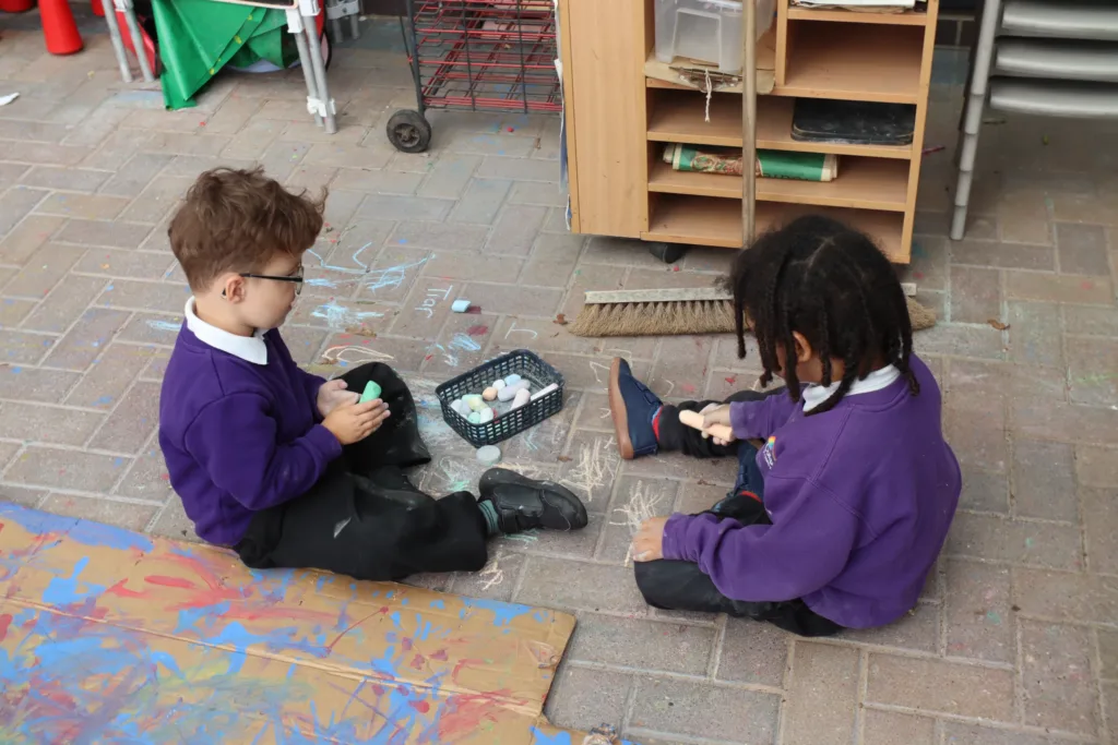 Two children in purple jumpers sit on the ground drawing with chalk. Art supplies and drawings surround them, and shelves with materials are in the background.