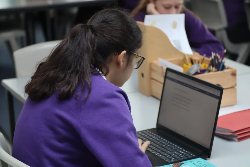 A student in a purple jumper works on a laptop at a desk in a classroom, with papers, pencils, and a book nearby. Another student is blurred in the background.