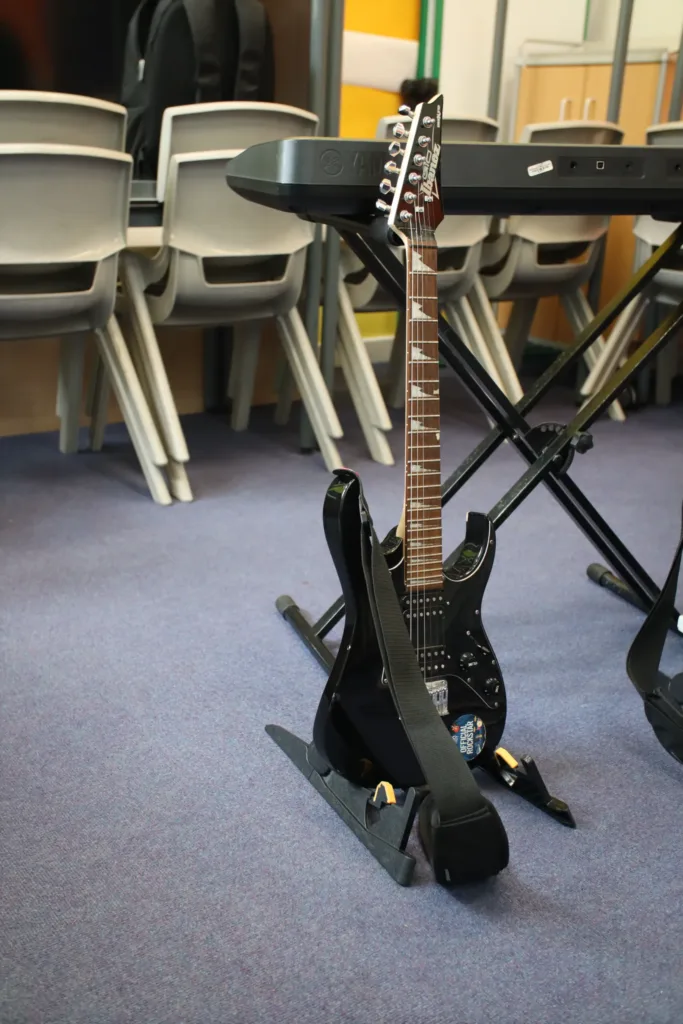 A black electric guitar with a strap rests on a stand beside an electronic keyboard in a classroom with stacked chairs in the background.