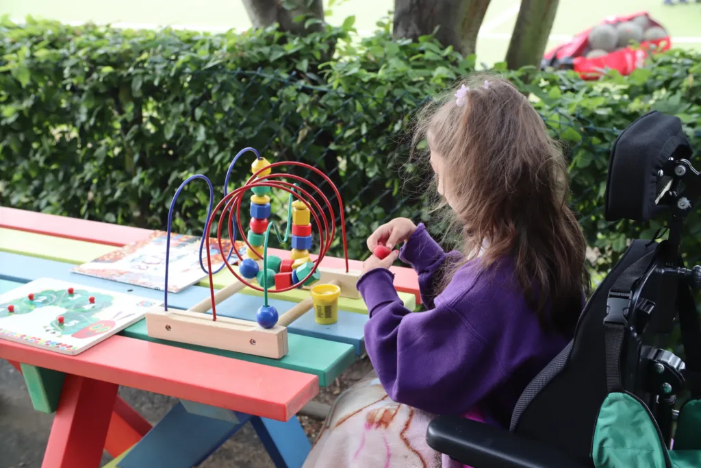 A girl in a wheelchair sits at a colourful outdoor table, playing with a bead maze and modelling clay. Trees and green bushes are in the background.
