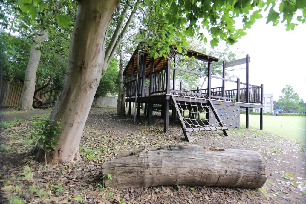 A wooden playground structure with a climbing net and slide stands among trees, with a large log in the foreground and leafy branches partially shading the scene.
