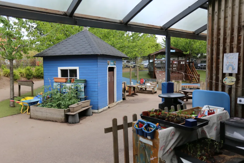 Outdoor play area with a blue playhouse, wooden tables, garden planters, a climbing frame, and children's toys and art supplies under a covered patio. Trees and parked cars are in the background.