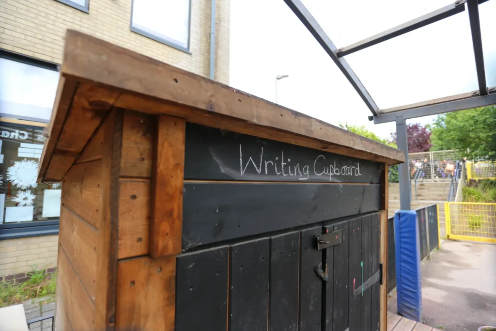 A wooden shed labelled Writing Cupboard in chalk sits outside a building near steps and a yellow fence.