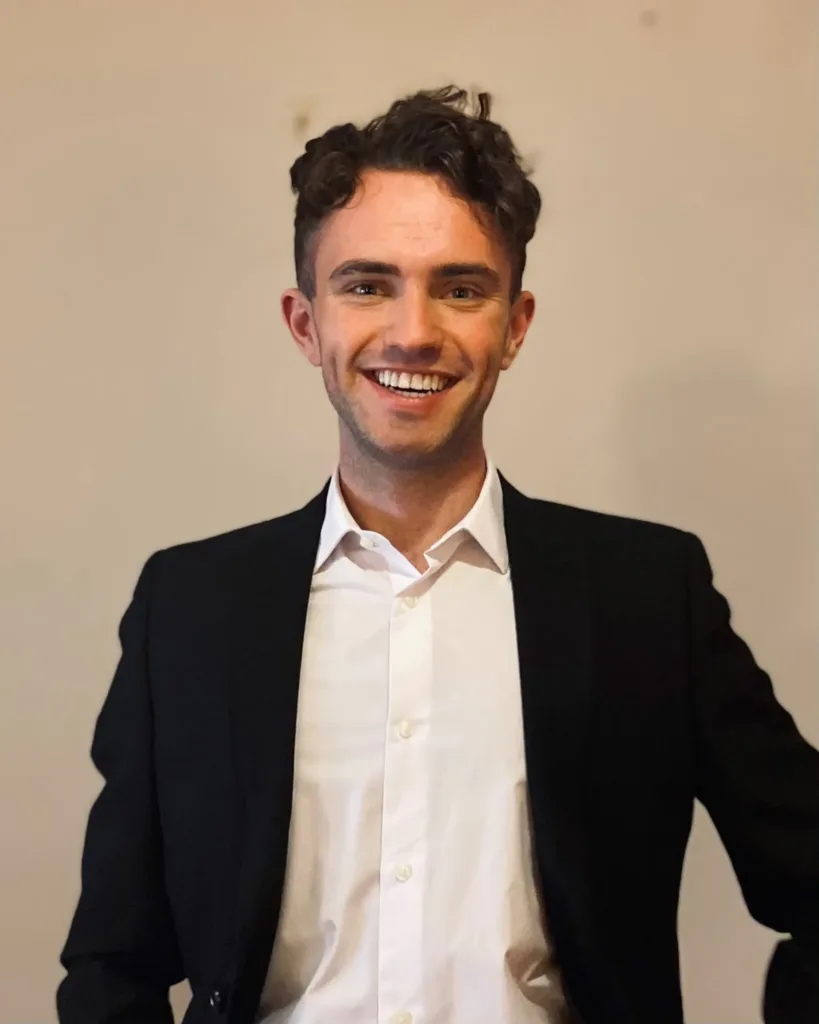 A young man with curly brown hair, wearing a black suit jacket and white shirt, smiles at the camera against a plain beige background.