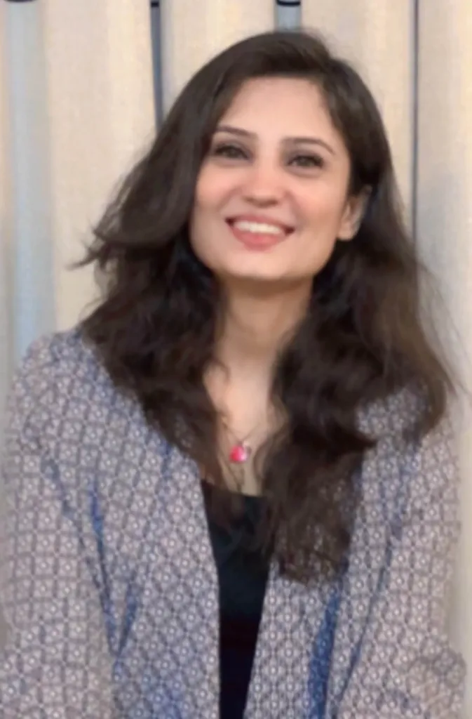 A woman with long, wavy dark hair smiles at the camera. She is wearing a patterned jacket over a black top with a pink pendant necklace. A light-coloured curtain is in the background.