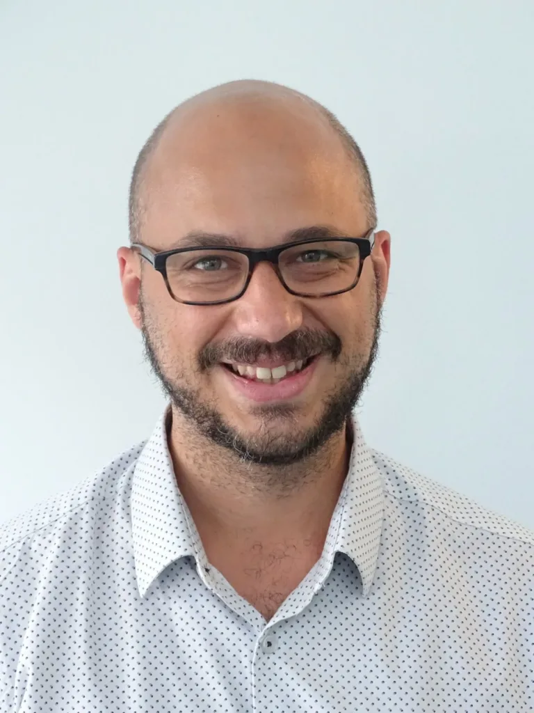 A smiling man with glasses, a bald head, and a short beard, wearing a white dress shirt with small black dots, standing in front of a plain light background.