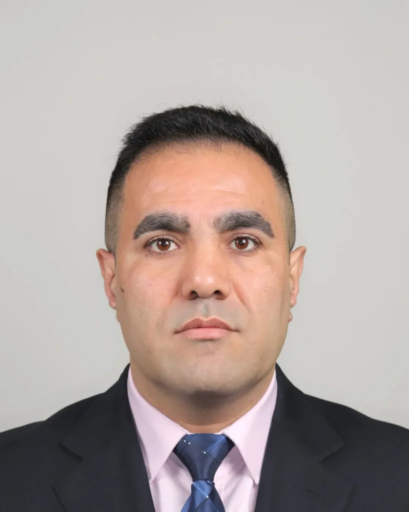 A man with short black hair, wearing a black suit, light pink shirt, and blue tie, is facing forward against a plain light grey background.